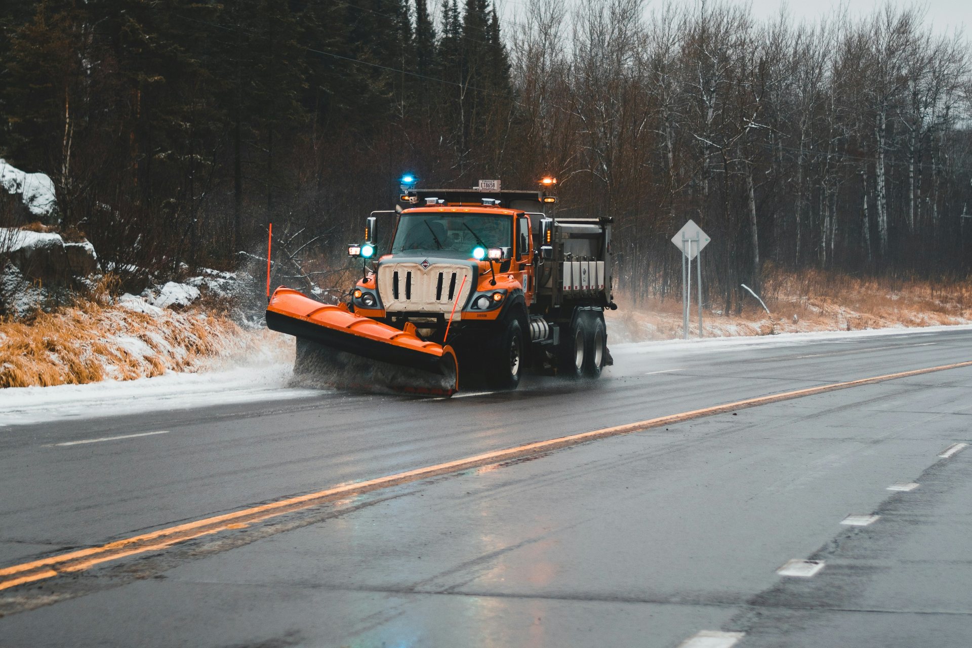 An orange plow truck clearing slush from a two-lane road lined with winter trees